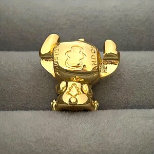 May include: A gold-colored charm featuring the character Stitch from Disney's Lilo & Stitch. The charm has Stitch's signature large ears and is engraved with the words "Disney" and "Pandora". The charm is made of a shiny, reflective material.
