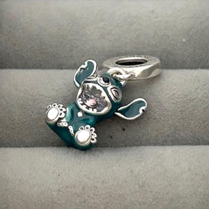 May include: A teal and silver charm featuring a cartoon character with large ears and a wide grin. The charm has a silver loop for attaching to a bracelet or necklace. The character's face is detailed with black and pink accents.