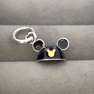 May include: A silver charm featuring a black and gold Mickey Mouse hat design. The charm has a silver loop for attaching to a bracelet or necklace. The hat has a gold Mickey Mouse head silhouette.