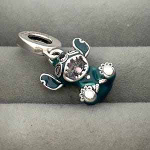 May include: A teal and silver charm in the shape of a cartoon character. The charm has a silver loop for attaching to a bracelet or necklace. The character has large ears, a round head, and a gem in the center of its face.