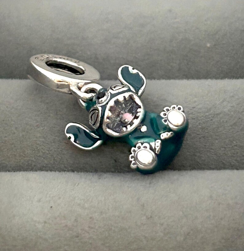 May include: A teal and silver charm in the shape of a cartoon character. The charm has a silver loop for attaching to a bracelet or necklace. The character has large ears, a round head, and a gem in the center of its face.
