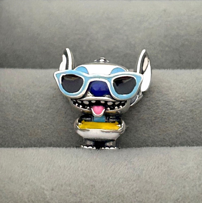 K&ouml;nnte beinhalten: Ein silberner Charm-Anh&auml;nger, der eine Zeichentrickfigur mit gro&szlig;en Ohren und blauer Sonnenbrille darstellt. Die Figur hat ein blaues Gesicht, eine rosa Zunge und ein gelb-wei&szlig;es Outfit. Dieser Charm ist wahrscheinlich f&uuml;r ein Armband oder eine Halskette.