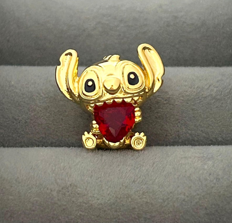 May include: A gold-colored Stitch figurine with a red heart-shaped gem. The figurine has large black eyes and raised ears. The gem is set in the center of the figurine's chest. The figurine is sitting on a gray surface.