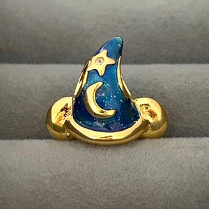 May include: A gold-colored wizard hat pin with a blue enamel finish. The hat features a gold crescent moon and star, with a small clear gem. The hat is set against a gray background.