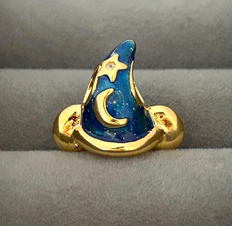 May include: A gold-colored wizard hat pin with a blue enamel finish. The hat features a gold crescent moon and star, with a small clear gem. The hat is set against a gray background.