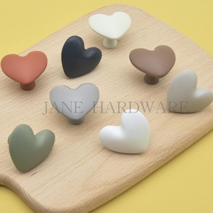 May include: A collection of heart-shaped cabinet knobs in various colors, including white, black, and muted shades of green, pink, and brown. The knobs are displayed on a wooden cutting board. The text "JANE HARDWARE" is visible.
