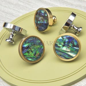 May include: A selection of decorative cabinet knobs featuring iridescent abalone shell inlays. The knobs have gold and silver finishes, with round and oval designs. The knobs are displayed on a light green surface.