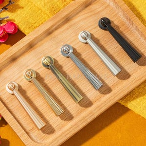 Può includere: Un vassoio di legno espone sei pomelli decorativi in varie finiture metalliche: oro, ottone, argento e nero. Ogni pomello presenta una testa cilindrica e un manico rettangolare scanalato. Sfondo giallo e fiori rossi.