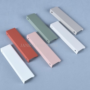 Könnte beinhalten: Eine Sammlung farbenfroher Schrankgriffe in verschiedenen Farbtönen, darunter Weiß, Rot, Rosa, Grün und Grau. Jeder Griff ist rechteckig mit einem gebogenen Griff und vorgebohrten Löchern. Der Text "JANE HARDWARE" ist sichtbar.