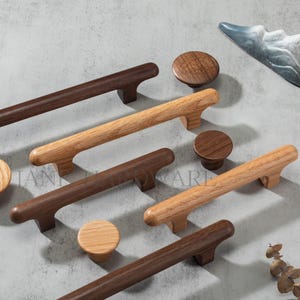 Può includere: Una collezione di ferramenta per mobili in legno, tra cui maniglie e pomelli in legno marrone scuro e chiaro. Le maniglie sono rettangolari con manici curvi e i pomelli sono rotondi. La ferramenta è disposta su una superficie grigia.