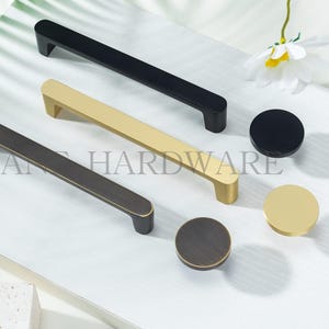 May include: A selection of cabinet hardware, including black, gold, and bronze-toned handles and knobs. The handles are rectangular, and the knobs are circular. The hardware is displayed on a white surface with a leaf and flower in the background.