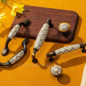 May include: Decorative cabinet hardware set featuring handles and knobs. The handles have a bronze finish with white and gold floral patterned ceramic inserts. The knobs have a matching design. The hardware is displayed on a wooden board.