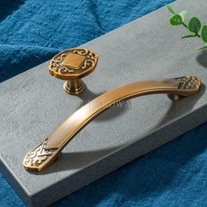 Könnte beinhalten: Antikes Messing-Schrankbeschlagsset. Enthält einen dekorativen Griff und einen passenden Knopf. Die Beschläge weisen aufwendige Details und eine warme, gealterte Oberfläche auf. Ideal, um Möbeln oder Schränken einen Vintage-Touch zu verleihen.