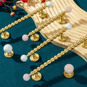 May include: A collection of gold-toned cabinet pulls and knobs. The pulls have a textured, wavy design with a white marble or pink quartz sphere at each end. The knobs have a similar design. The hardware is displayed on a wooden surface.