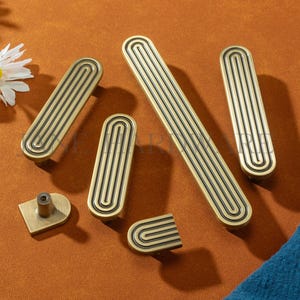 Peut inclure: Ensemble de quincaillerie de meubles en laiton antique, comprenant des poignées et un bouton. Les poignées sont rectangulaires avec un motif rainuré. L'ensemble est présenté sur une surface brun-orangé.