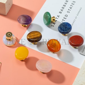 May include: A collection of colorful gemstone cabinet knobs with gold-toned bases. The knobs feature various stones, including amethyst, tiger's eye, and rose quartz. The knobs are arranged on a white surface with text and a peach-colored background.