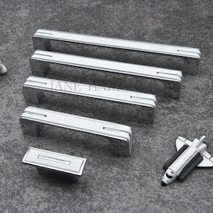 May include: A collection of silver-toned cabinet pulls in various sizes, with a central recessed panel. The pulls are arranged with a small astronaut figurine and a toy space shuttle. The pulls have a modern, streamlined design.