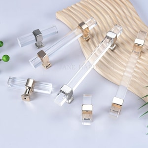 Może przedstawiać: Przezroczyste akrylowe uchwyty do szafek ze złotymi i srebrnymi metalowymi akcentami. Uchwyty są prostokątne i mają nowoczesny design. Obraz zawiera kilka uchwytów o różnych rozmiarach, ułożonych na jasnej powierzchni.