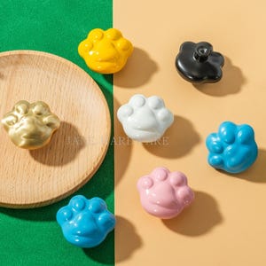 May include: A collection of paw-shaped cabinet knobs in various colors, including gold, yellow, white, black, blue, and pink. The knobs are arranged on a green and tan surface, with a wooden plate visible. The text "JANE HARDWARE" is also visible.