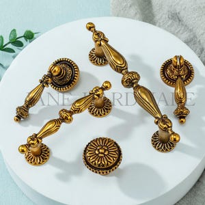 May include: A collection of antique brass cabinet hardware, including handles and knobs. The hardware features ornate detailing and a warm, aged finish. The pieces are arranged on a white surface, showcasing their decorative design.