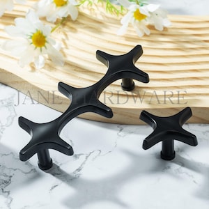 Może przedstawiać: Czarne okucia do szafek w kształcie gwiazdy. Na zdjęciu widać trzy elementy okuć, w tym długi uchwyt i dwa gałki. Widoczny jest tekst "JANE HARDWARE". Okucia są prezentowane na drewnianej desce z białymi kwiatami.