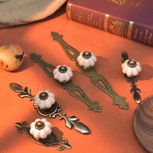 May include: A collection of antique-style drawer pulls and knobs. The pulls have a bronze finish with white ceramic flower-shaped knobs. The knobs are arranged on an orange surface with a wooden bird and a book in the background.