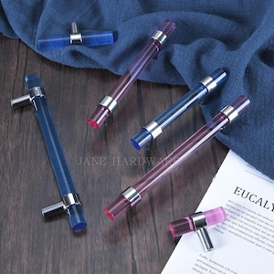 May include: A collection of colorful acrylic cabinet pulls in shades of blue and pink, with silver-toned metal accents. The pulls are arranged on a dark wood surface with a blue fabric backdrop. The text "JANE HARDWARE" is visible.