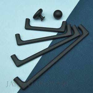 May include: A selection of matte black cabinet hardware, including pull handles, a knob, and a round pull. The hardware is displayed on a two-tone blue background. The brand name is visible in the lower left corner.