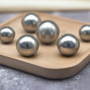 Puede incluir: Seis esferas metálicas plateadas y brillantes de varios tamaños están dispuestas sobre una bandeja de madera marrón claro. Las esferas reflejan la luz, creando reflejos y sombras. La bandeja tiene un borde liso y curvado.