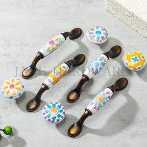 Puede incluir: Un conjunto de herrajes decorativos para armarios, que incluye tiradores y pomos a juego. Los tiradores tienen un acabado en bronce y centros de cerámica blanca con diseños florales coloridos en rosa, azul, amarillo y naranja. Los pomos tienen diseños similares.