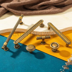 Peut inclure: Un assortiment de quincaillerie d'armoire en laiton antique, comprenant des poignées et des boutons. La quincaillerie présente divers motifs texturés, notamment un motif en forme de corde, des rainures linéaires et une texture pointillée. Les pièces sont disposées sur une surface en bois.