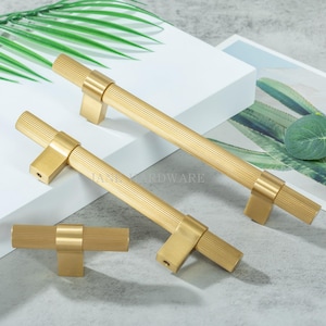 May include: Three gold-colored cabinet pulls with a ribbed design. The pulls are rectangular with a T-bar shape. The image is well-lit, showing the hardware against a neutral background. The text "JAMES HARDWARE" is visible.