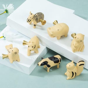 Puede incluir: Un conjunto de tiradores de armario con forma de animales, incluyendo una cebra, un hipopótamo y una ardilla. Cada tirador es de color beige claro con detalles negros y un asa dorada. Se presentan sobre una superficie blanca.