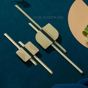 Könnte beinhalten: Zwei messingfarbene Schrankgriffe mit modernem Design. Jeder Griff hat einen rechteckigen Mittelteil mit gerilltem oder geriffeltem Muster, der mit langen, schlanken Stäben verbunden ist. Der Text "JANE HARDWARE" ist sichtbar.