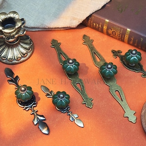 May include: An assortment of antique-style cabinet hardware, including drawer pulls and knobs. The hardware features a dark green, pumpkin-shaped ceramic knob with a bronze-colored metal base. The drawer pulls have an elongated design with decorative accents.