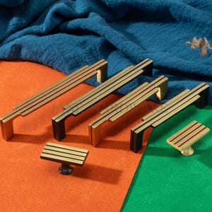 May include: A collection of gold-toned cabinet hardware, including handles and a knob, with black accents. The hardware features a linear design with three parallel lines. The items are displayed on a colorful orange and green surface.