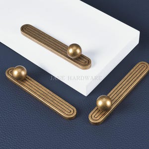May include: Three antique brass cabinet pulls. Each pull features a rectangular base with grooved detailing and a spherical knob. The pulls are arranged on a white surface against a dark blue background. The text "JANE HARDWARE" is visible.