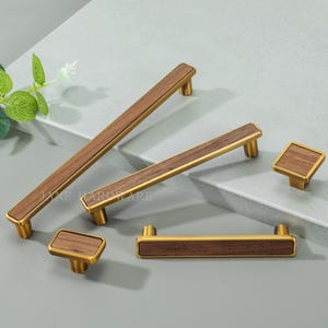 May include: A collection of cabinet hardware featuring a warm wood grain center with a gold-toned metal frame. The set includes various handle sizes and square knobs, offering a cohesive design for furniture upgrades.