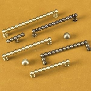 May include: An assortment of antique-style cabinet pulls and knobs in various sizes and finishes. The hardware features a beaded design and is available in brass and bronze tones. The image is set against a warm, neutral background.