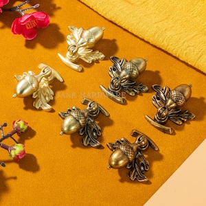 May include: Decorative acorn-shaped cabinet knobs in gold and bronze tones. The knobs feature intricate leaf and acorn designs, with a curved base for easy installation. The image shows several knobs arranged on a yellow surface, suggesting a hardware or home decor theme.