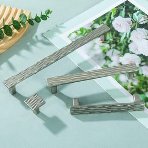 May include: A set of silver-coloured cabinet handles and a square knob with a textured, wavy design. The items are displayed on a light blue surface, with a floral print in the background. The handles are suitable for kitchen or furniture use.