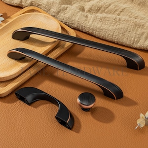 Op de afbeelding: Een set zwarte kastbeslagen met koperkleurige accenten. De set bevat twee lange handgrepen, een kleinere handgreep en een ronde knop. De hardware wordt gepresenteerd op een bruin, leerachtig oppervlak.