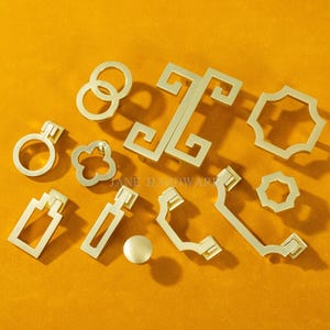 May include: A collection of gold-colored cabinet hardware, including handles, knobs, and pulls, arranged on a mustard-yellow surface. The hardware features various geometric and floral designs, with the text "JANE HARDWARE" visible.