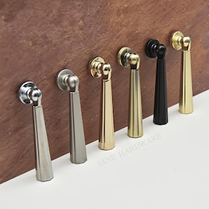 May include: A collection of six cabinet pulls in various metallic finishes, including silver, gold, and black. Each pull features a circular base and a tapered, elongated handle. The pulls are arranged against a brown surface.