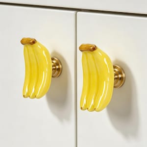 May include: Two cabinet knobs shaped like a bunch of yellow bananas with brown tips. Each banana-shaped handle is attached to a circular, brushed gold-toned metal base, mounted onto a smooth, off-white cabinet door surface for functional use.