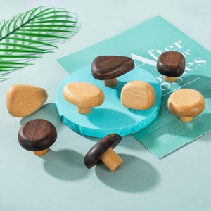 May include: A collection of wooden cabinet knobs in various shapes and shades of brown and tan. The knobs are displayed on a light blue surface, with a few green leaves in the background. The knobs have a mushroom-like design.