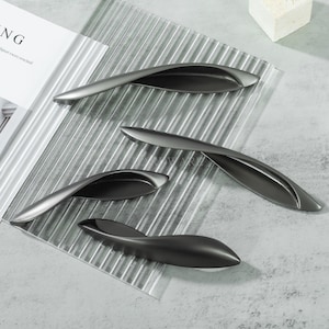 Puede incluir: Una taza de cerámica blanca con un asa negra y el texto "But First, Coffee" en negrita negra. La taza se encuentra sobre un fondo borroso, sugiriendo un ambiente acogedor.