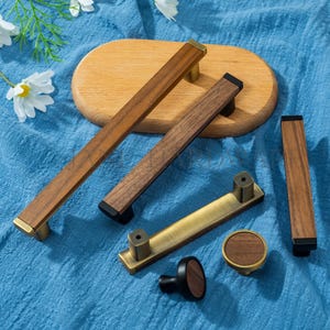 May include: A selection of wooden cabinet handles and knobs. The handles are rectangular with brass or black detailing. The knobs have a round wooden face. The hardware is displayed on a blue fabric surface. Measurements are in centimetres.