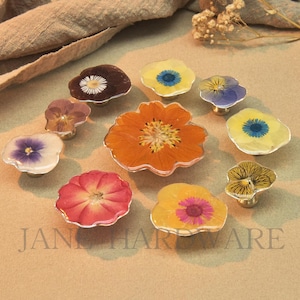 May include: A collection of floral cabinet knobs with various pressed flower designs. The knobs feature a clear resin finish with silver-colored metal bases. The flowers include orange, yellow, red, purple, and white blooms. The text "JANE HARDWARE" is visible.