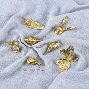 May include: A selection of gold-coloured decorative hardware, including a leaf-shaped pull, a rabbit head, a seashell, a butterfly, and bird-shaped pulls. The hardware is displayed on a grey fabric surface. The text "JANE HARDWARE" is visible.